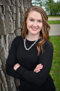 Photo of Dr. Julie Stamm. Julie has her hair down and is crossing her arms leaning up against a tree. She is in a black shirt wearing a necklace and there is grass and paved blacktop in the background.