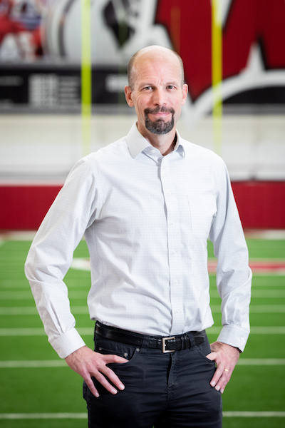 Dr. Peter Miller standing portrait. Peter is in a white shirt with his hands at his side. He is standing on grass with a motion W for the Wisconsin Badgers in the background. The yellow goal post can also be seen in the background.