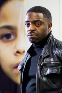 Dr. Faisal Abdu'Allah portrait. Faisal is wearing a black turtleneck with a black leather jacket. He is in the forefront of the photo and behind him is a photograph of a half face of a young child.