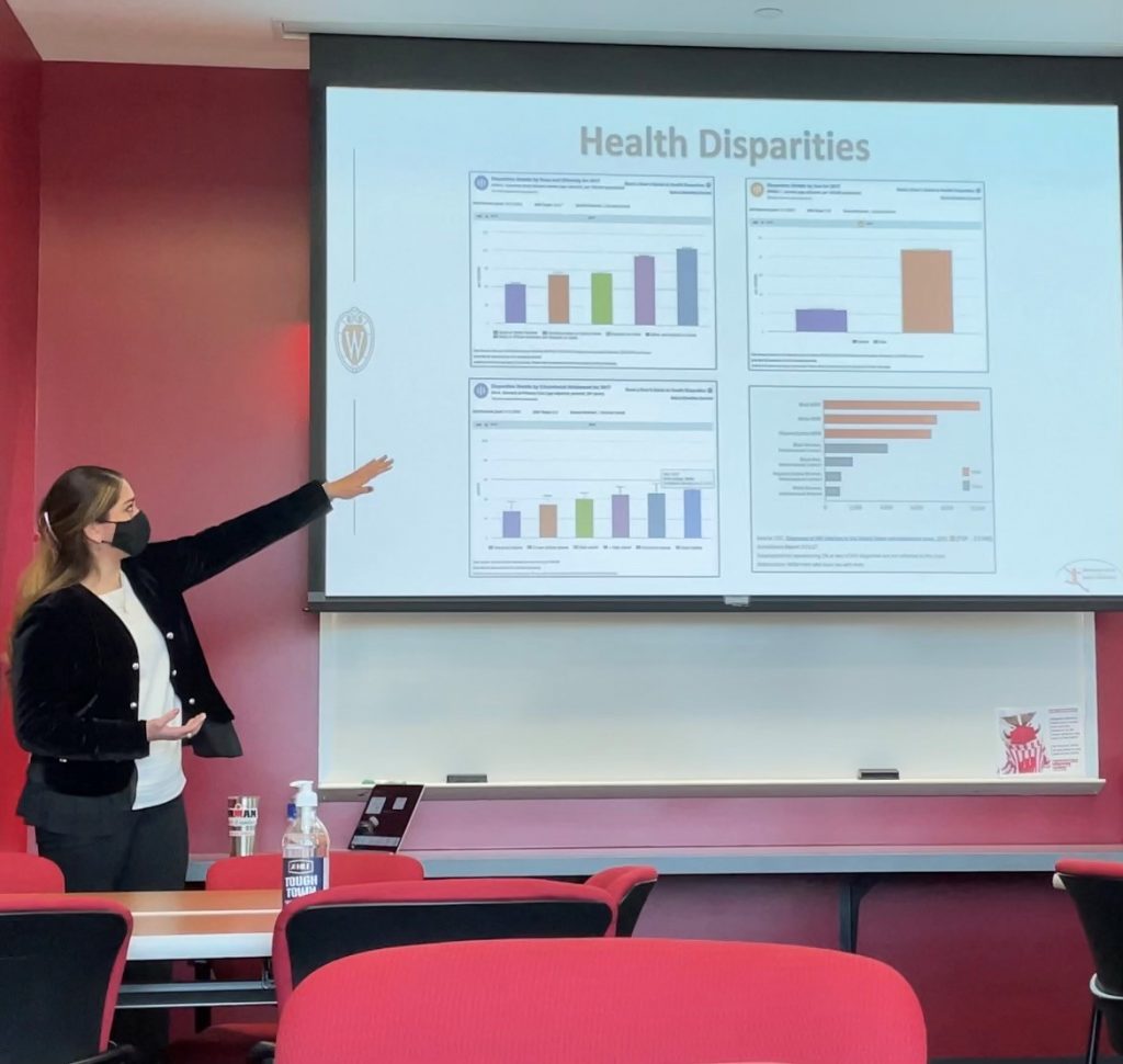 A photo of a woman wearing a mask. She is presenting a powerpoint. The picture shows the PowerPoint in the middle of the screen and the woman is on the left of the screen looking at the screen with her arm raised toward the screen. 