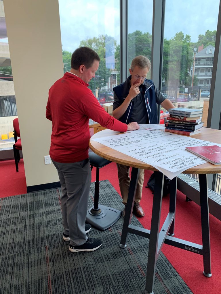 Two men are standing at a table looking at a sheet of paper. One man in red is pointing to the paper while the other man in black has his hand at his mouth, pondering the sheet of paper. There are large poster sheets on the table and there is a stack of books on the table. In the background, there are floor to ceiling windows and out of the windows, there are houses. 