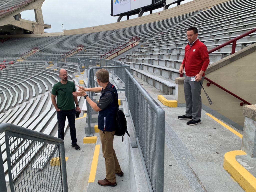 Picture of three men in the stands at Camp Randall Stadium. One man is standing on the top step, wearing a red shirt. One man is standing on the middle step, wearing a black shirt. One man is on the bottom step, wearing a green shirt. The man in the black shirt is talking with his hands clapped together. The stadium is empty bleachers. 