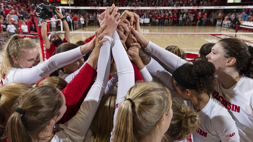 UW Badger volleyball team huddle together raising up their arms together. The volleyball court and volleyball net are in the background. The team is wearing white long-sleeve shirts.