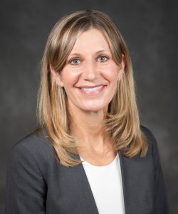 Posed head shot of Dr. Ann Sheehy. It is a gray background and Ann is centered in the photo. She is wearing a white shirt with a gray blazer.