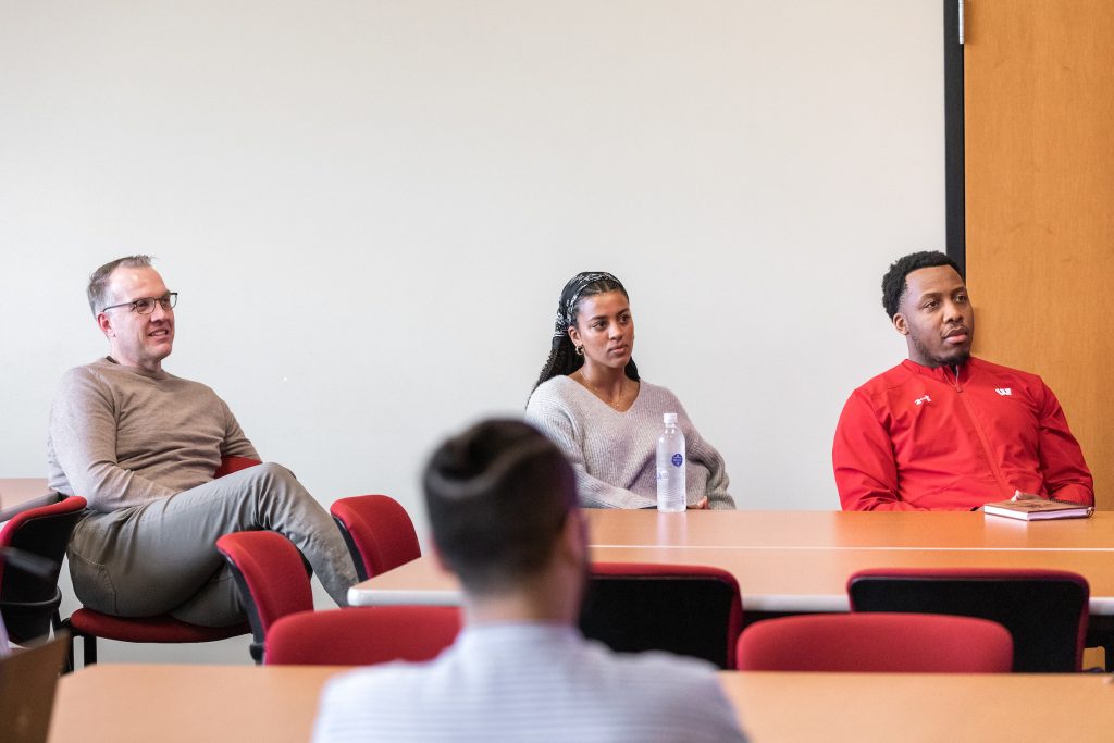Pictured are three people sitting down listening to the presentation. There is a table in front of the people watching the presentation. Unfocused is the backside of someone sitting down. 