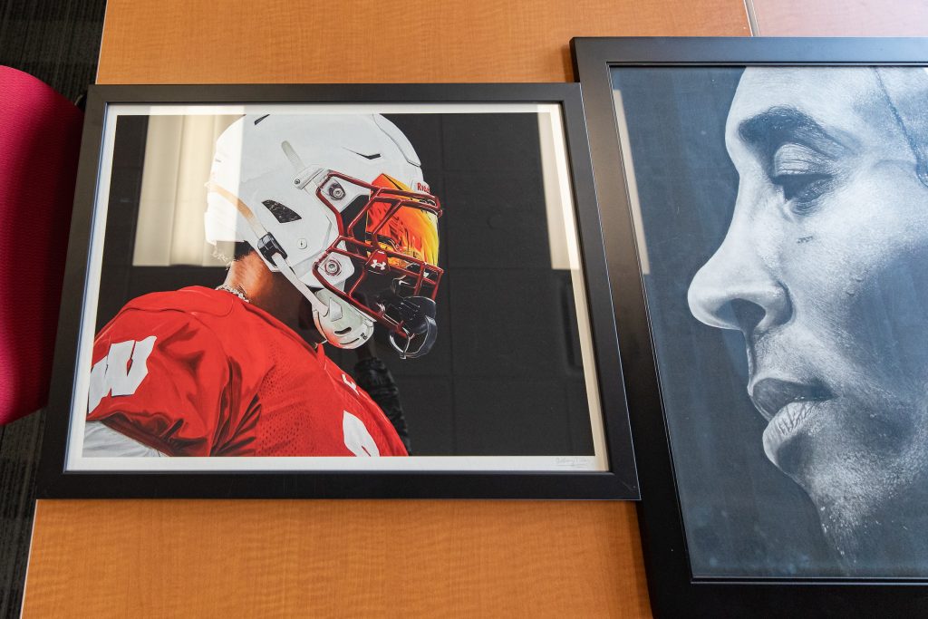 Two photos are on a table. One photo is of a football player in a Badgers jersey and is wearing a helmet. The helmet has an eye screen that is shaded red and orange. The second phot is a charcoal drawing of Kobe Bryant. 