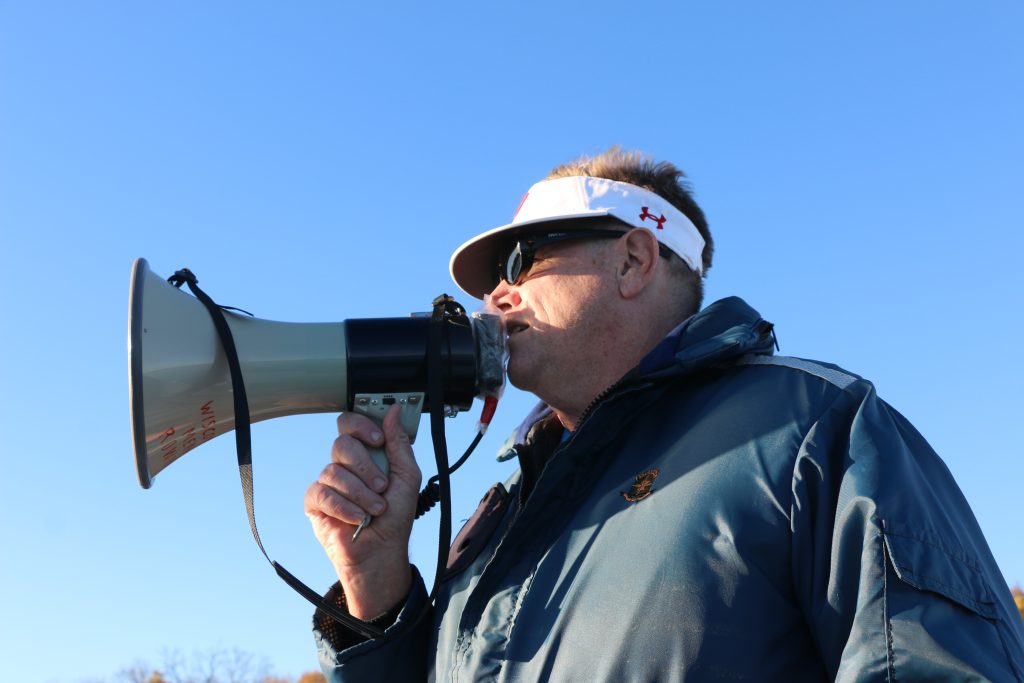 Chris Clark is wearing a blue jacket and a white visor that has an Under Armour logo on the side. He is speaking into a megaphone that he is holding. He is outside and the sky is blue.