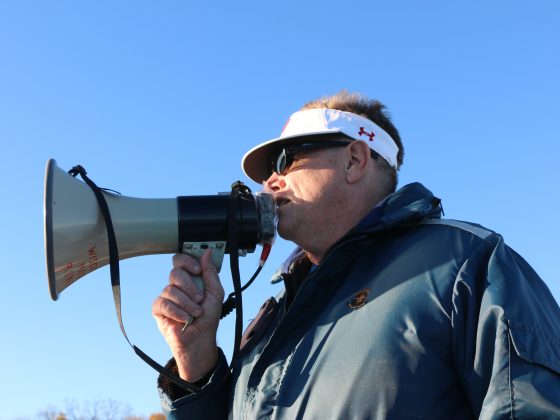 Chris Clark is wearing a blue jacket and a white visor that has an Under Armour logo on the side. He is speaking into a megaphone that he is holding. He is outside and the sky is blue.