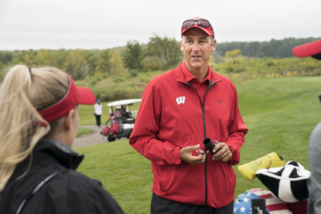 Women's Golf Head Coach, Todd Oehrlein, is wearing a red jacket with a red hat. He is speaking to a player who is wearing a black jacket and a red visor. He is holding a range finder and they are on a golf course. The grass is green and there's brush in the background. There are golf clubs next to Todd.