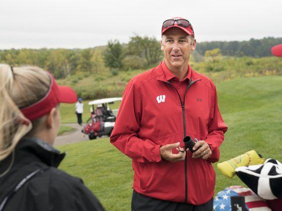 Women's Golf Head Coach, Todd Oehrlein, is wearing a red jacket with a red hat. He is speaking to a player who is wearing a black jacket and a red visor. He is holding a range finder and they are on a golf course. The grass is green and there's brush in the background. There are golf clubs next to Todd.
