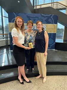 Kathy is standing in the middle with Josie to her left and a former Baraboo High School teacher to her right. Josie is holding a bell cow in front of Kathy. They are standing in front of stage smiling for the camera.