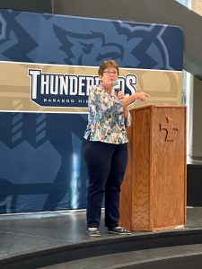 Kathy Allen stands on a stage next to a wooden podium. She is holding a microphone and pointing to the crowd. She is giving a speech. Behind her is a Thunderbirds Baraboo High School Sign.