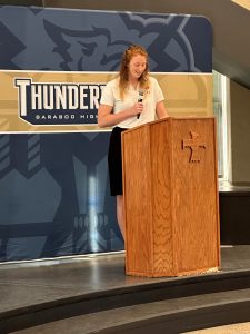 Josie is holding a microphone while standing behind a wooden podium on a stage. She is giving a speech to the crowd.