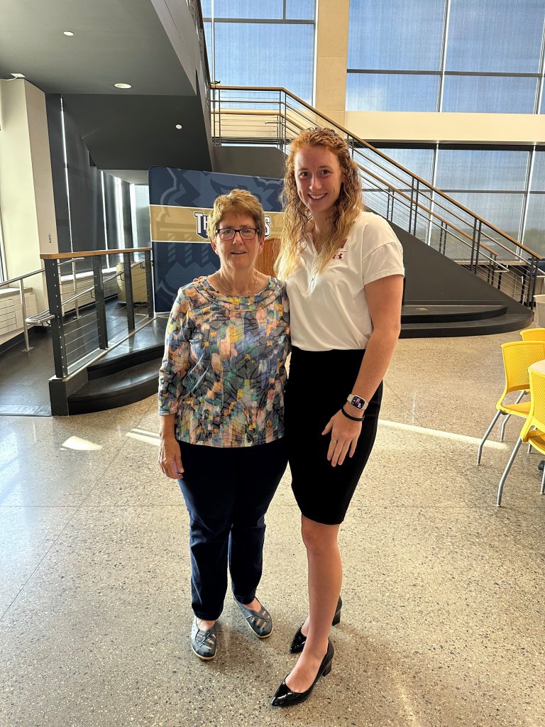 Josie and Kathy pose for a picture. Josie is wearing a white shirt and a black skirt. Kathy is to her left wearing a floral shirt and jeans.