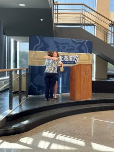 Josie Schaefer embraces Kathy Allen in a hug while standing on the podium. Josie is holding the bell cow award and is standing next to a podium.