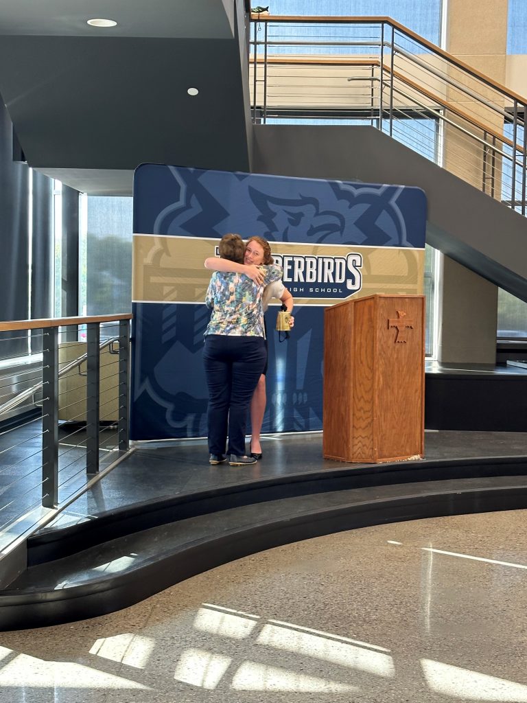 Josie Schaefer embraces Kathy Allen in a hug while standing on the podium. Josie is holding the bell cow award and is standing next to a podium.