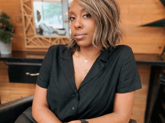Pictured is Shayla Yon. She is wearing a black shirt and is seated. She is leaning her elbow on a chair. She is sitting down at table. There is a mirror behind her.