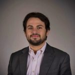 Landry Levinson Professional Headshot Pictured is a man in a gray suit jacket and purple collard shirt. He has dark hair and facial hair.