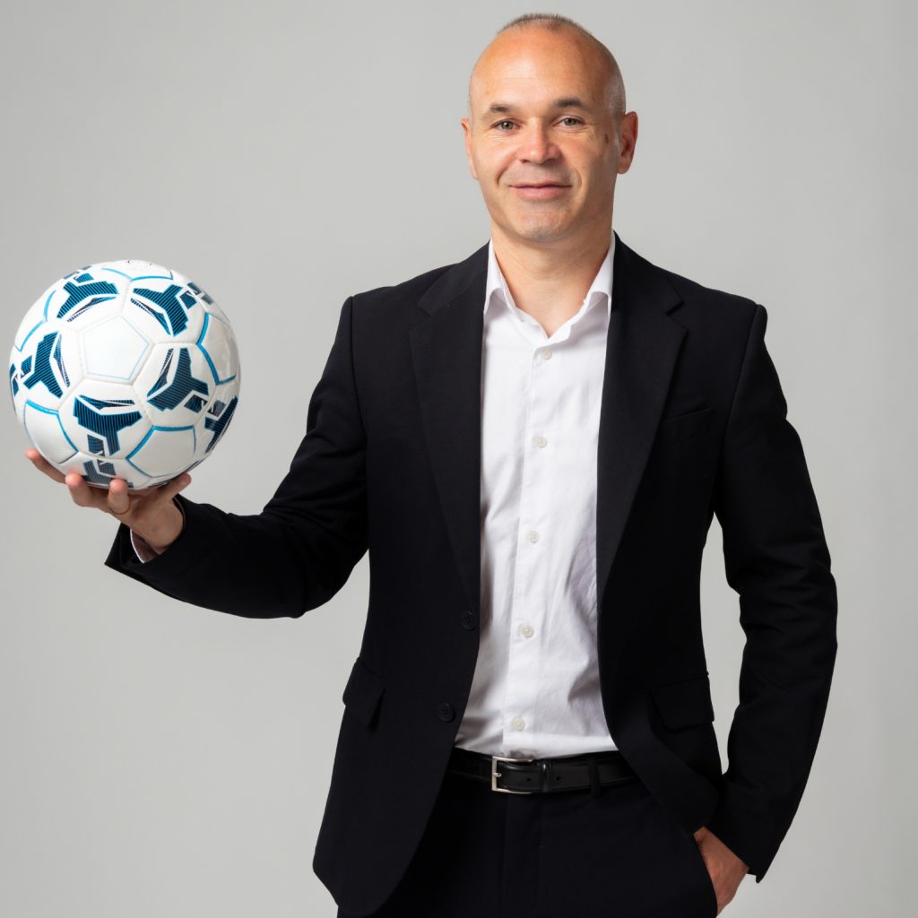 Posed is Andres Iniesta. He is facing the camera standing in a black suit jacket and a white shirt. He is holding out a soccer ball with his right hand. He is smiling.