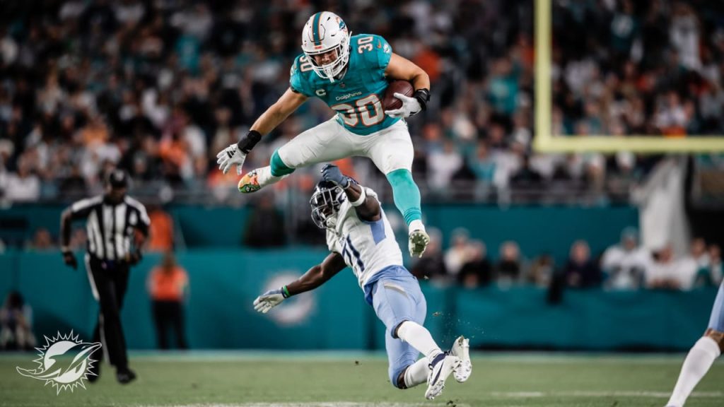 Pictured is Alec Ingold, a football player for the Miami Dolphins. He is jumping over an opponent while holding a football. A referee stands in the background with fans in the stands. Photo curtsey of the Miami Dolphins.