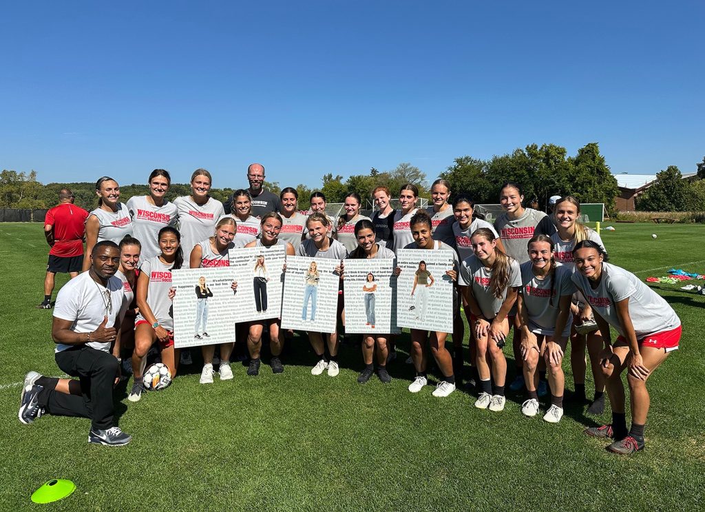 Pictured is the Wisconsin Women's soccer team with professor Faisal Abdu'Allah. They are standing on green grass and are lined up in two rows, with the first row squating. Five players in the first row are holding printed photos of their teammates in street clothes.