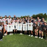 Pictured is the Wisconsin Women's soccer team with professor Faisal Abdu'Allah. They are standing on green grass and are lined up in two rows, with the first row squating. Five players in the first row are holding printed photos of their teammates in street clothes.