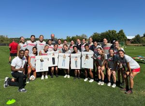 Pictured is the Wisconsin Women's soccer team with professor Faisal Abdu'Allah. They are standing on green grass and are lined up in two rows, with the first row squating. Five players in the first row are holding printed photos of their teammates in street clothes.
