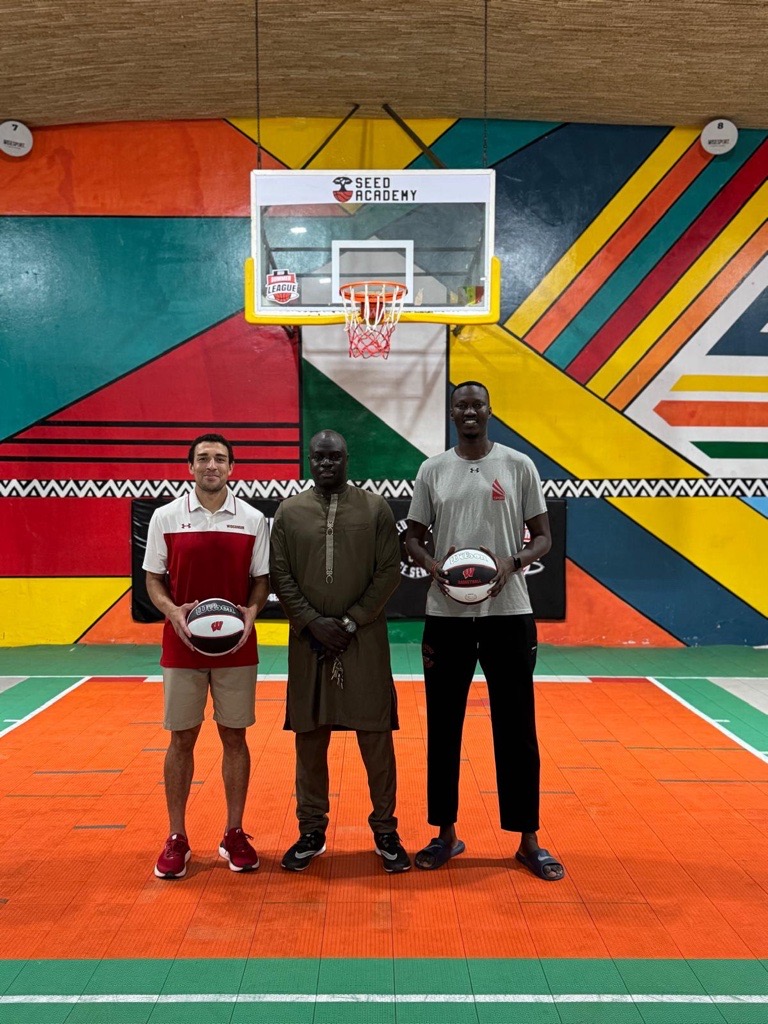 Pictured are three people in the center of the court in front of a basketball hoop. The floor is orange and green and the color on the wall is multicolored. The three men are standing and smiling. The two men on the outsides are holding basketballs and the man in the center has his hands crossed against his middle.