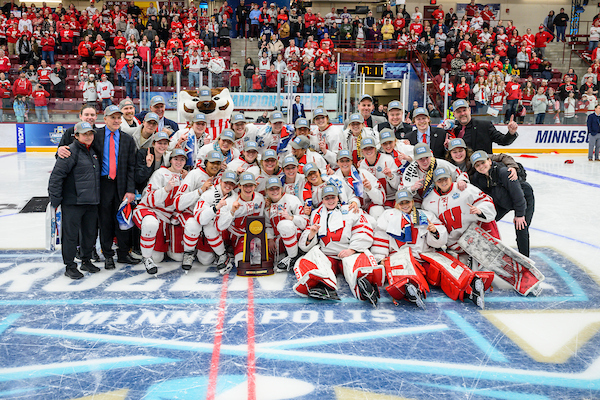 Pictured center ice is the Wisconsin women's hockey team with the coaches, players, and bucky. They are wearing ball caps and holding a trophy.