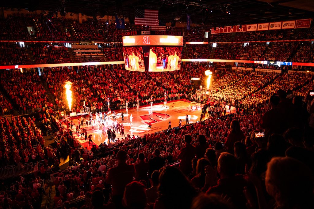 Pictured is a basketball court with the lights off. The court is lit by fire, showcasing players on the court and fans in the stands. It is a wide angle shot showing three levels of a full stadium.