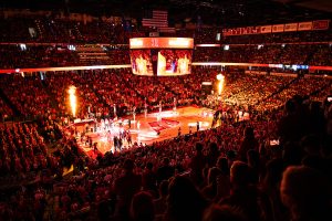 Pictured is a basketball court with the lights off. The court is lit by fire, showcasing players on the court and fans in the stands. It is a wide angle shot showing three levels of a full stadium.