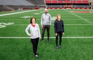 Three people stand on a football field inside Camp Randall Stadium. From left, Maria Dehnert, Peter Miller, and Sara Jimenez Soffa pose for a photo.