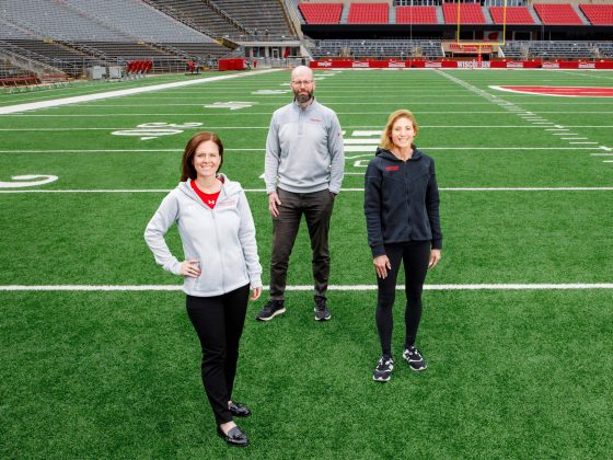 Three people stand on a football field inside Camp Randall Stadium. From left, Maria Dehnert, Peter Miller, and Sara Jimenez Soffa pose for a photo.