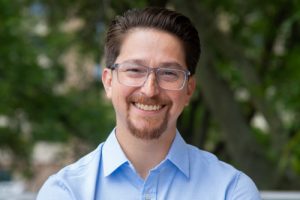 Professional outdoor headshot of Dr. Jason Kido Lopez smiling, wearing glasses, a goatee, and a light blue button-down shirt, with blurred green trees in the background.