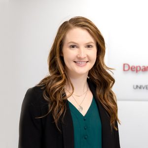 Professional headshot of Dr. Julie Stamm smiling, wearing a black blazer over a teal blouse, with a blurred university department sign in the background.