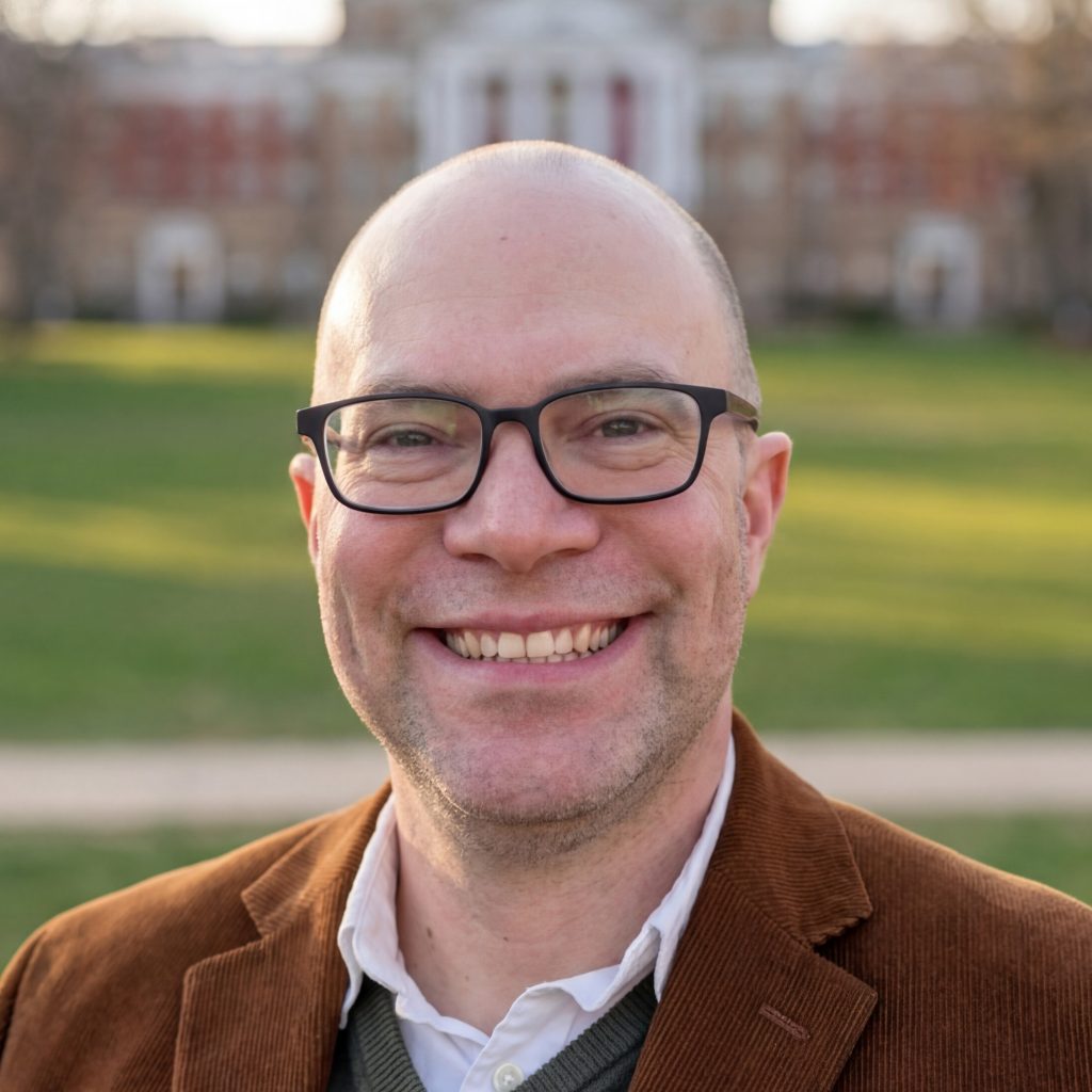 Headshot of Michael McCann smiling outdoors on a university campus, wearing a brown corduroy blazer and glasses, with a brick building and green lawn visible in the background.