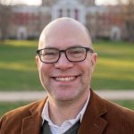 Mike Portrait Headshot of Michael McCann smiling outdoors on a university campus, wearing a brown corduroy blazer and glasses, with a brick building and green lawn visible in the background.