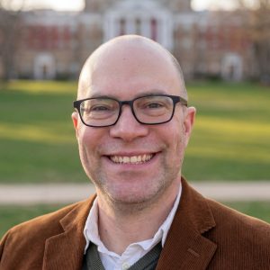 Headshot of Michael McCann smiling outdoors on a university campus, wearing a brown corduroy blazer and glasses, with a brick building and green lawn visible in the background.