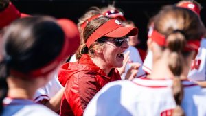 Yvette Healy, head coach of the Wisconsin Badgers softball team, energetically addresses her players in a dugout huddle during a game. She is wearing a red Wisconsin visor, sunglasses, and a red vest, surrounded by players in white and red Badgers uniforms.
