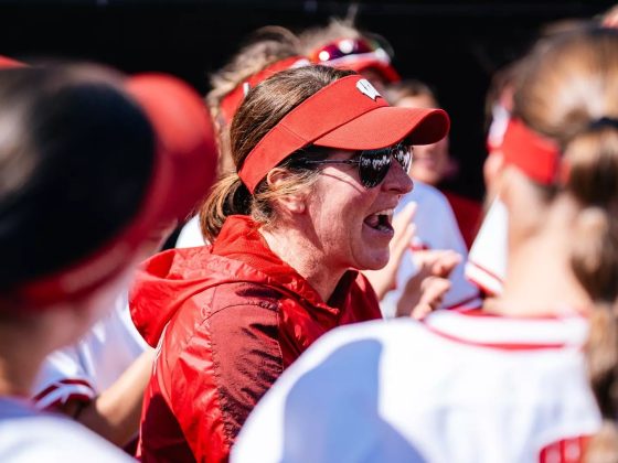 Yvette Healy, head coach of the Wisconsin Badgers softball team, energetically addresses her players in a dugout huddle during a game. She is wearing a red Wisconsin visor, sunglasses, and a red vest, surrounded by players in white and red Badgers uniforms.