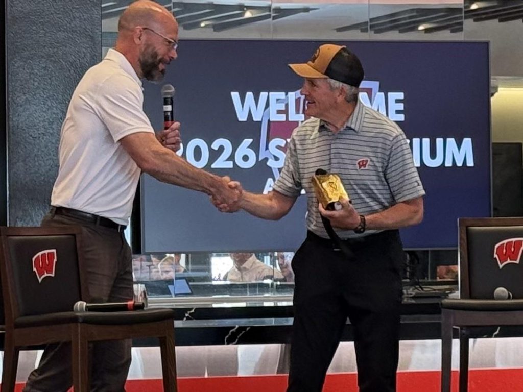 Bell Cow Award Dr. Peter Miller shakes hands with UW hockey coach Mark Johnson as Johnson holds the Bell Cow Award during the 2026 symposium.