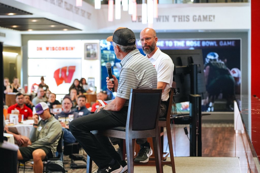 DSC08366 Large UW hockey coach Mark Johnson speaks into a microphone while Dr. Peter Miller listens during a symposium panel at Camp Randall Stadium.