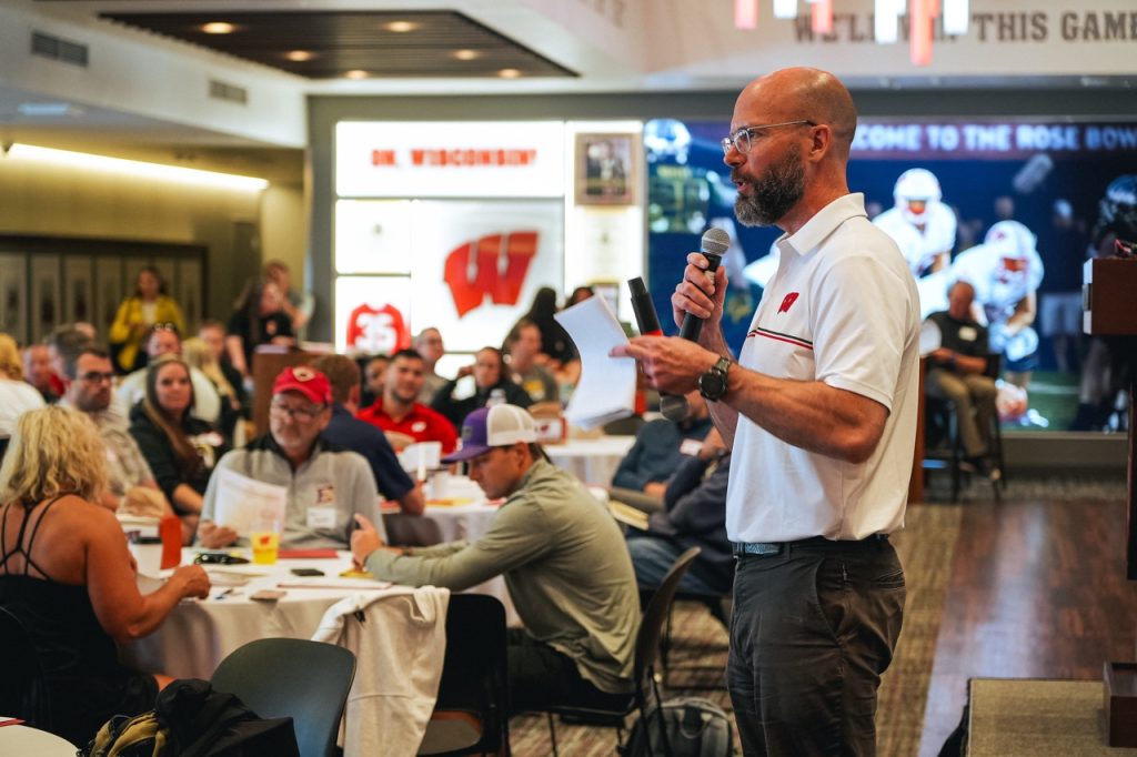 DSC08863 Large BIOS Director Peter Miller addresses Symposium participants while holding a microphone and notes inside Camp Randall Stadium.