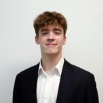 Ben Schweitzer Headshot of Ben Schweitzer, a smiling young man with wavy brown hair wearing a black suit jacket and a white open-collared shirt against a plain white background.