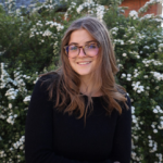 Lindsey Cholewczynski Portrait of Lindsey Cholewczynski, a smiling young woman with long brown hair and glasses, wearing a black long-sleeved top in front of a green bush with white flowers.